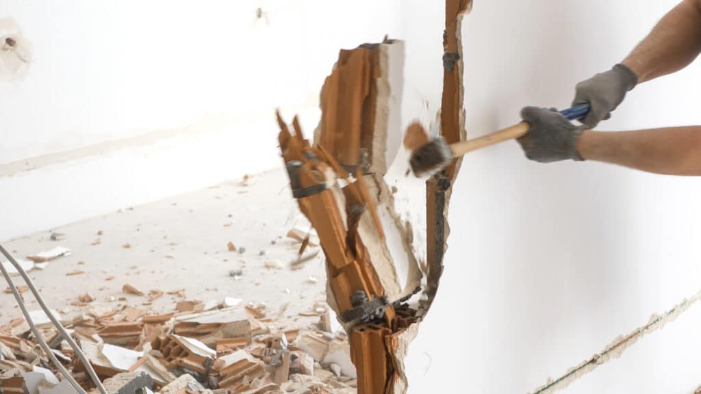 Démolition mur briques : ouvrier utilise une masse Ouvrier avec gants cassant un mur intérieur en briques creuses avec une masse, débris au sol.